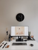 A minimalist desk setup features a sleek monitor displaying a fashion website. On the desk are accessories like a wristwatch, AirPods, a lamp, a bottle, a candle, a plant, and books, arranged neatly. Above the desk is an artistic clock with numbers clustered on one side.