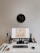A minimalist desk setup features a sleek monitor displaying a fashion website. On the desk are accessories like a wristwatch, AirPods, a lamp, a bottle, a candle, a plant, and books, arranged neatly. Above the desk is an artistic clock with numbers clustered on one side.