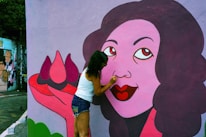 A person wearing a white tank top and denim shorts is painting the details on a colorful mural that depicts a woman with purple hair and vibrant red lips, holding a large flower. The mural is set against an outdoor urban backdrop with other pieces of street art visible.