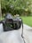 A camera with a large lens is placed on a surface outdoors. The camera's screen displays various settings and a small part of a scene being captured, potentially part of a garden or natural area. The background is filled with green grass and dense leafy trees, creating a lush and serene atmosphere.