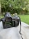 A camera with a large lens is placed on a surface outdoors. The camera's screen displays various settings and a small part of a scene being captured, potentially part of a garden or natural area. The background is filled with green grass and dense leafy trees, creating a lush and serene atmosphere.
