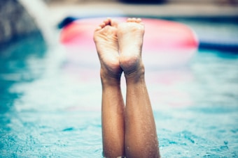 Legs are raised vertically out of the water in a swimming pool, with droplets of water visible on the skin. In the background, there's an out-of-focus inflatable pool ring.
