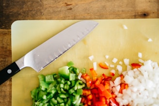 A cutting board displaying chopped green peppers, red peppers, and onions alongside a knife with a black handle. The board rests on a wooden surface.