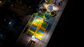 A colorful, illuminated amusement ride with trampolines and bungee cords viewed from above. The bungee trampolines are arranged in a circular formation, each with brightly colored rings in yellow, green, and blue. Surrounding the structure is a darkened outdoor space with some vegetation and pathways visible.