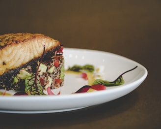 A gourmet dish featuring a fillet of grilled fish, possibly salmon, atop a bed of quinoa and vegetables, garnished with microgreens and delicate red leaves, served on a white plate.