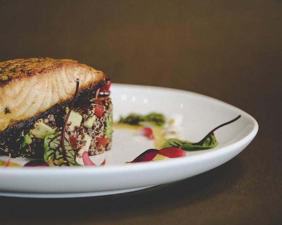 A gourmet dish featuring a fillet of grilled fish, possibly salmon, atop a bed of quinoa and vegetables, garnished with microgreens and delicate red leaves, served on a white plate.