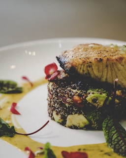 A close-up view of a gourmet dish plated elegantly. The dish features a piece of grilled fish on top of a bed of quinoa mixed with diced vegetables and garnished with vibrant edible flowers and fresh herbs. A creamy sauce is artfully drizzled around the components.