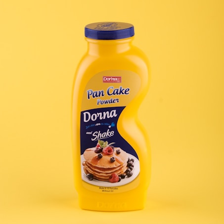 A yellow plastic bottle of pancake powder stands against a matching yellow background. The label is blue and displays the brand name, 'Dorna,' along with an image of stacked pancakes garnished with berries and mint leaves. The label also includes the text 'Pan Cake Powder' and 'Shake' and indicates the mix makes 8-10 pancakes without oil.