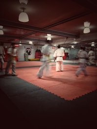 A martial arts dojo with several people practicing. The room is well-lit with ceiling lights and a soft red-pink mat covers the floor. Participants are dressed in traditional martial arts uniforms, focusing on their movements.