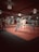 A martial arts dojo with several people practicing. The room is well-lit with ceiling lights and a soft red-pink mat covers the floor. Participants are dressed in traditional martial arts uniforms, focusing on their movements.