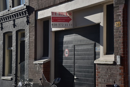 A street scene featuring a dark-colored building with a garage door. Above the door, there is a 'Te Huur' sign indicating availability for rent, with contact information included. The building has a mix of brickwork textures in shades of grey and brown. An indistinct object, possibly part of a motorcycle, is visible in the foreground.
