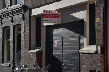 A street scene featuring a dark-colored building with a garage door. Above the door, there is a 'Te Huur' sign indicating availability for rent, with contact information included. The building has a mix of brickwork textures in shades of grey and brown. An indistinct object, possibly part of a motorcycle, is visible in the foreground.