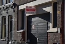 A street scene featuring a dark-colored building with a garage door. Above the door, there is a 'Te Huur' sign indicating availability for rent, with contact information included. The building has a mix of brickwork textures in shades of grey and brown. An indistinct object, possibly part of a motorcycle, is visible in the foreground.