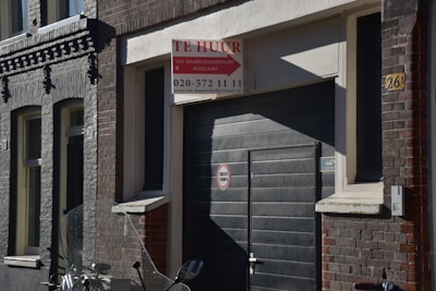 A street scene featuring a dark-colored building with a garage door. Above the door, there is a 'Te Huur' sign indicating availability for rent, with contact information included. The building has a mix of brickwork textures in shades of grey and brown. An indistinct object, possibly part of a motorcycle, is visible in the foreground.