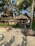 A tropical beach scene featuring several thatched huts surrounded by lush green palm trees. The sandy beach has multiple lounge chairs facing away from the ocean, suggesting a relaxing vacation spot.
