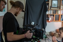 A group of people are involved in a filmmaking or video production setting. One person is holding a professional camera setup, complete with a monitor and various accessories. They appear focused on the camera's screen. In the background, there is a black backdrop, a bookshelf, and some lighting equipment.