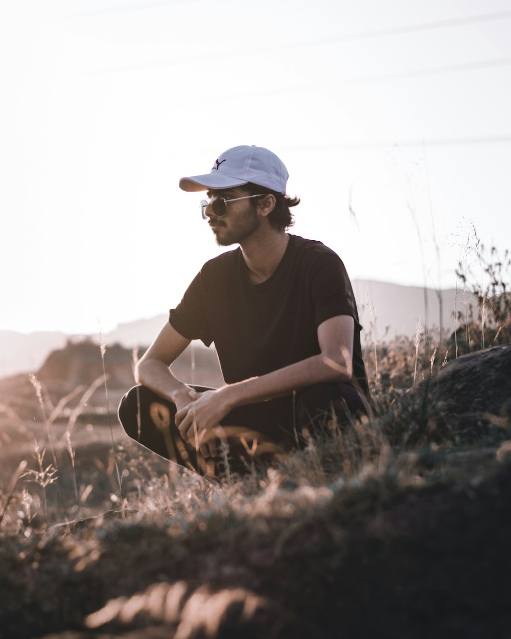 man in black crew neck t-shirt and white cap sitting on brown grass during daytime