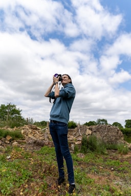 A person is standing outdoors in a natural setting, holding a camera to take photos. The individual is dressed in casual clothing and appears focused on capturing images. The background includes some greenery and scattered rocks, along with a partly cloudy sky above.