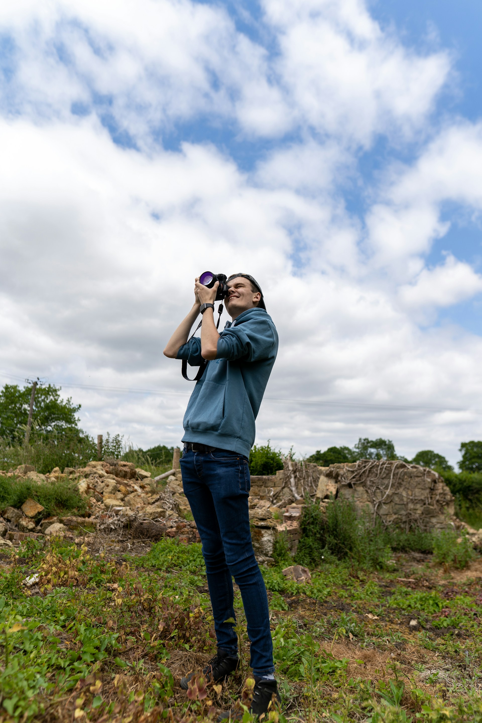 A person is standing outdoors in a natural setting, holding a camera to take photos. The individual is dressed in casual clothing and appears focused on capturing images. The background includes some greenery and scattered rocks, along with a partly cloudy sky above.