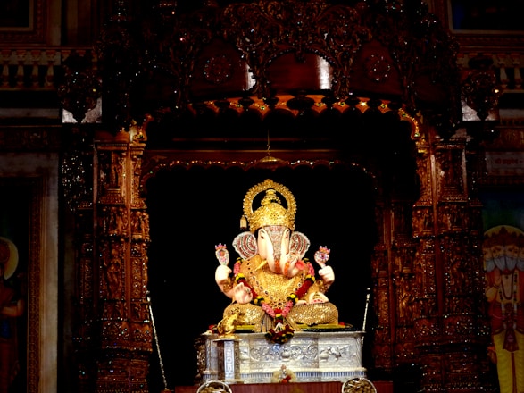 A golden statue of the Hindu deity Ganesha is intricately decorated and placed on a silver pedestal inside a richly carved wooden shrine. Strings of flowers and other ornate details surround the figure, which is illuminated against the darker backdrop.