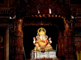 A golden statue of the Hindu deity Ganesha is intricately decorated and placed on a silver pedestal inside a richly carved wooden shrine. Strings of flowers and other ornate details surround the figure, which is illuminated against the darker backdrop.