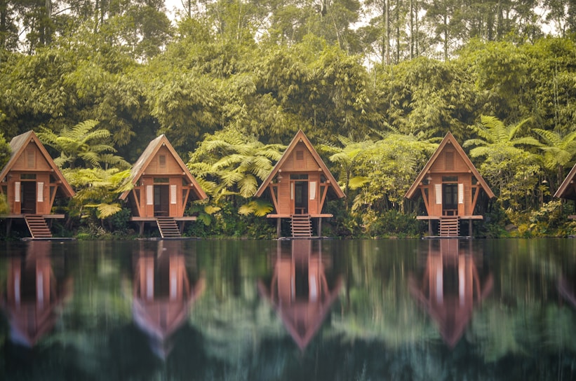 A row of wooden A-frame cabins is nestled among lush greenery and tall trees. Each cabin stands on stilts above a serene body of water, with clear reflections visible on the surface. The scene is surrounded by dense vegetation, including large ferns and bamboo, creating a tranquil and secluded atmosphere.