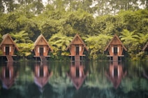 A row of wooden A-frame cabins is nestled among lush greenery and tall trees. Each cabin stands on stilts above a serene body of water, with clear reflections visible on the surface. The scene is surrounded by dense vegetation, including large ferns and bamboo, creating a tranquil and secluded atmosphere.