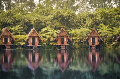 A row of wooden A-frame cabins is nestled among lush greenery and tall trees. Each cabin stands on stilts above a serene body of water, with clear reflections visible on the surface. The scene is surrounded by dense vegetation, including large ferns and bamboo, creating a tranquil and secluded atmosphere.