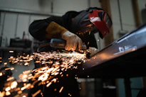 A person wearing a welding helmet grinding a metal beam with sparks flying