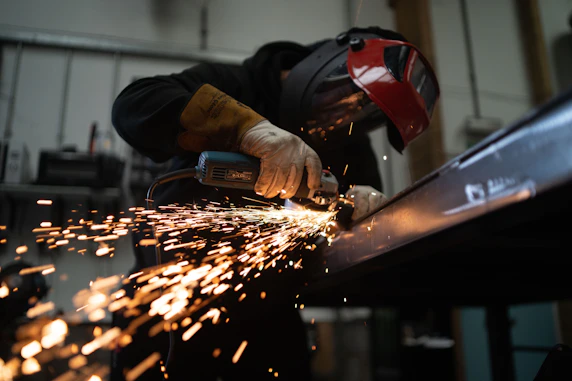 A person wearing a welding helmet grinding a metal beam with sparks flying