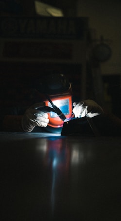 A person is engaged in welding, wearing a protective helmet and gloves. The scene is dimly lit, with the bright glow of the welding arc illuminating the hands and tools. The focus is on the work being done, with a concentration of light and shadows.