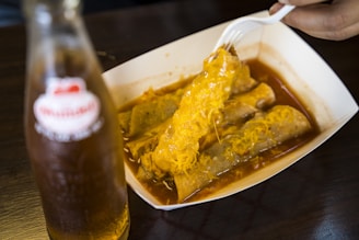 A plate containing rolled tacos covered in melted cheese and sauce, with a hand holding a fork lifting one of the tacos. A bottle of soda is placed to the side.