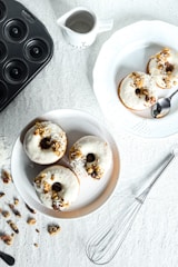 On a textured white surface, three doughnuts are arranged on a round plate, topped with white icing, nuts, and coconut flakes. A spoon rests on a separate plate with another doughnut. A whisk, scattered nuts, and a black donut pan are arranged nearby, alongside a small pitcher.