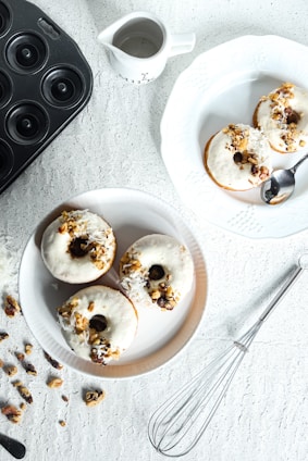 On a textured white surface, three doughnuts are arranged on a round plate, topped with white icing, nuts, and coconut flakes. A spoon rests on a separate plate with another doughnut. A whisk, scattered nuts, and a black donut pan are arranged nearby, alongside a small pitcher.