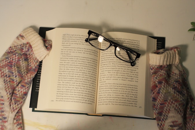 An open book with pages displaying text, accompanied by reading glasses placed on top. The book is flanked by a pair of colorful knitted socks, suggesting a cozy reading environment. The surface is light-colored with a small green plant visible in the corner.