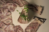 A folded book stands on a surface with green leaves sprouting from it, surrounded by a warm, patterned sweater. A pair of black glasses rests next to the book. The setting appears cozy and artistic, blending natural elements with a sense of homeliness.