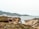 A rocky coastal landscape with rugged cliffs and a few people exploring the area. The sea is calm, and the sky is overcast. Vegetation covers parts of the foreground with earthy and green tones, while the background features distant hills.