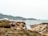 A rocky coastal landscape with rugged cliffs and a few people exploring the area. The sea is calm, and the sky is overcast. Vegetation covers parts of the foreground with earthy and green tones, while the background features distant hills.