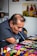 A man with a brush in his mouth, painting vibrant art pieces at a table filled with colorful crafts. He is focused on his work, displaying intricate designs and vivid colors. The background shows artwork with people and sunflowers.