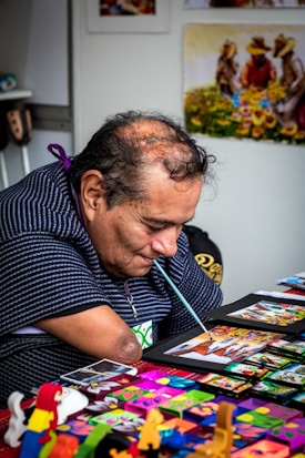 A man with a brush in his mouth, painting vibrant art pieces at a table filled with colorful crafts. He is focused on his work, displaying intricate designs and vivid colors. The background shows artwork with people and sunflowers.