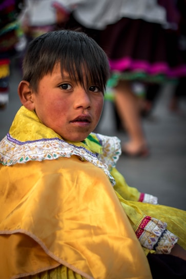 A young boy with dark hair gazes directly at the camera. He is dressed in a vibrant yellow traditional outfit with intricate embroidery and lace details around the collar. The background is slightly blurred, showing other people dressed in colorful attire.