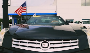 A black Cadillac is prominently displayed with a reflection of clouds on its hood. An American flag is visible in the background, indicating perhaps a dealership setting. There is a yellow sticker on the windshield showing a year, suggesting the car is for sale. A part of a white building and another car are also visible.