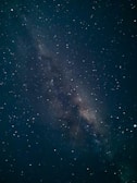 A stunning view of the Milky Way galaxy with countless stars scattered across the night sky. The center of the galaxy is visible, creating a dense, luminous band stretching diagonally. A faint shooting star is visible, adding an element of motion to the serene scene.