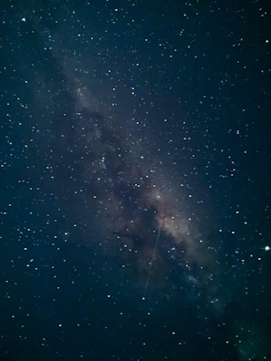 A stunning view of the Milky Way galaxy with countless stars scattered across the night sky. The center of the galaxy is visible, creating a dense, luminous band stretching diagonally. A faint shooting star is visible, adding an element of motion to the serene scene.