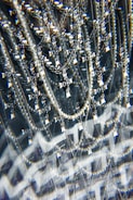 A complex assemblage of sparkling jewelry featuring multiple chains and a prominent cross pendant. The scene is visually intensified by a kaleidoscopic effect, adding layers of optical distortion and reflections. The jewelry hangs against a dark background, enhancing the contrast and highlighting the metallic and pearl-like textures.
