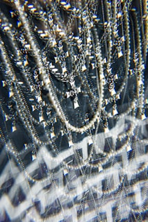 A complex assemblage of sparkling jewelry featuring multiple chains and a prominent cross pendant. The scene is visually intensified by a kaleidoscopic effect, adding layers of optical distortion and reflections. The jewelry hangs against a dark background, enhancing the contrast and highlighting the metallic and pearl-like textures.