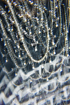 A complex assemblage of sparkling jewelry featuring multiple chains and a prominent cross pendant. The scene is visually intensified by a kaleidoscopic effect, adding layers of optical distortion and reflections. The jewelry hangs against a dark background, enhancing the contrast and highlighting the metallic and pearl-like textures.