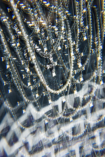 A complex assemblage of sparkling jewelry featuring multiple chains and a prominent cross pendant. The scene is visually intensified by a kaleidoscopic effect, adding layers of optical distortion and reflections. The jewelry hangs against a dark background, enhancing the contrast and highlighting the metallic and pearl-like textures.
