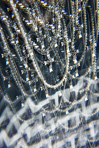 A complex assemblage of sparkling jewelry featuring multiple chains and a prominent cross pendant. The scene is visually intensified by a kaleidoscopic effect, adding layers of optical distortion and reflections. The jewelry hangs against a dark background, enhancing the contrast and highlighting the metallic and pearl-like textures.