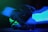 A motorcycle helmet is placed against a dark backdrop with vibrant blue and green light streaks creating an ethereal effect. The helmet is sleek with a glossy finish, and its patterns are highlighted by the neon lights.